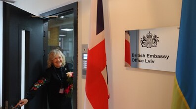 Усміхнена жінка відчиняє двері офісу, поруч табличка з написом «British Embassy Office Lviv» та прапори Великої Британії й України