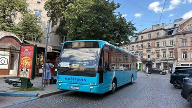 Міський автобус бірюзового кольору маршруту №67 стоїть на брукованій дорозі біля зупинки громадського транспорту. На тлі видно старовинну забудову, зелені дерева та кілька припаркованих автомобілів.