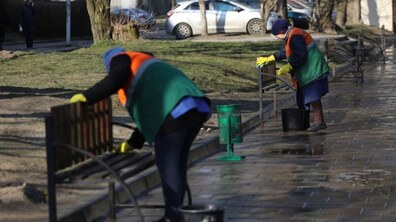 Дві працівниці комунальної служби прибирають територію вздовж тротуару. Вони у зелених жилетах із помаранчевими світловідбивними елементами та жовтих рукавицях миють лавки водою з відер. Навколо видно дерева, припарковані автомобілі та вологу бруківку, що блищить на сонці.