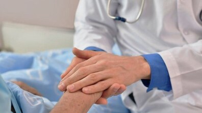 Лікар у білому халаті тримає руку пацієнта: видно лише частину тіл – руки та фрагмент одягу. Пацієнт лежить на ліжку, накритий світло-блакитною ковдрою. Жест виглядає турботливим і співчутливим: лікар накрив долонею руку пацієнта, ніби заспокоюючи чи підтримуючи його