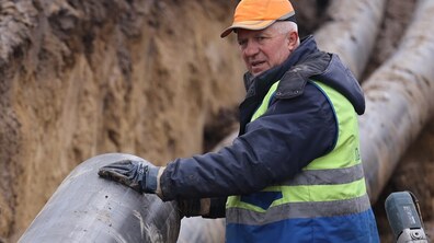 Чоловік середнього віку, стоїть у глибокій траншеї з ґрунтовими стінами. На чоловікові помаранчева кепка, темна куртка та яскравий світловідбивний жилет поверх синього робочого одягу. На руках — робочі рукавиці. Перед ним — велика пластикова труба великого діаметра; чоловік тримає її руками, ніби вирівнює або оглядає місце з’єднання. Позаду видно ще кілька таких самих труб, покладених уздовж траншеї.