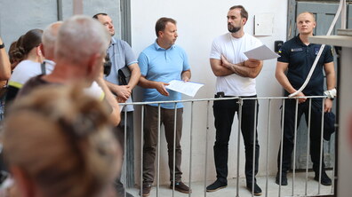 Представники міста та працівники Державної служби надзвичайних ситуацій стоять на вузькому балконі старого будинку та слухають один одного. У руках в них документи.
