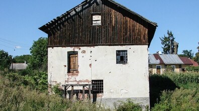 На фото зображено старовинний двоповерховий водяний млин із пошкодженим фасадом, оточений травою та приватною забудовою