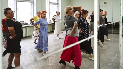 На фото група жінок займається танцями у світлій залі перед великим дзеркалом. Учасниці повторюють рухи, тримаючи руки на поясі