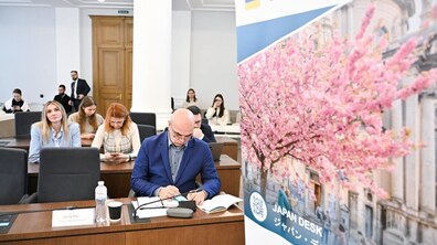 люди сидять в залі, слухають і занотовують у записники. На передньому плані видно банер з сакурою. Фото ілюстративне.