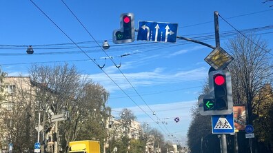 Перехрестя зі світлофорами, де увімкнене червоне світло та додаткова зелена стрілка ліворуч. Поруч розташовані дорожні знаки напрямків руху по смугах, головної дороги, а також пішохідного переходу й велосипедного переїзду. На проїзній частині їдуть легкові авто темного кольору та жовта вантажівка.