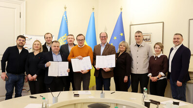 По центру стоїть Андрій Москаленко, перший заступник міського голови Львова, з права від нього Айке Фатер (Eike Vater), керівник проєкту «EMPOWER», а зліва Олександр Одинець, директор ЛМКП «Львівтеплоенерго». Усі троє тримають відкриті папки з підписаним меморандумом. Позаду і поруч з ними стоять депутати Львівської міської ради та представники Німецького товариства GIZ