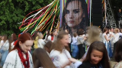 Сцена під час фестивалю ЧЕКАFEST у Шевченківському Гаю. На передньому плані багато людей – переважно молодь у традиційному українському вбранні з вишиванками та червоними намистами. Рух людей створює ефект розмиття, що надає знімку динаміки. На задньому плані – велика інсталяція з вертикальними кольоровими стрічками та портретом загиблої бойової медикині, громадської діячки та Героїні України Ірини Цибух.