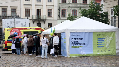 Білий медичний намет з написом «Перевір своє здоров’я» на площі Ринок у Львові. Біля намету зібралася черга людей, які прийшли на безкоштовні обстеження. Поруч стоїть автомобіль швидкої допомоги, а на самому наметі є інформаційні матеріали про здоровий спосіб життя.