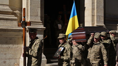 Похорон захисника. Військовослужбовці у формі несуть хрест, портрет, труну та синьо-жовтий Прапор України. Позаду мури Гарнізонного храму св. ап. Петра та Павла
