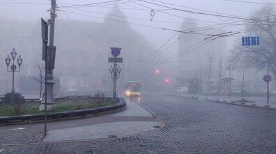 міська вулиця, вкрита бруківкою, огорнута густим туманом. Попереду дорогою їде жовтий тролейбус із увімкненими фарами. Видно дорожні знаки та ліхтарі. Силуети будівель і світлофори ледве проглядаються крізь сіру імлу.