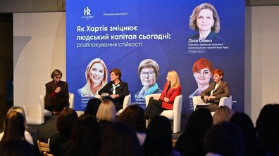 На сцені сидять четверо жінок. Одна з них тримає у ркуах мікрофон. Позаду на великому екрані розміщені їхні обличчя та напис білими літерам – Як Хартія зміцнює людський капітал сьогодні. На них із залу дивляться глядачі