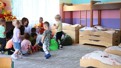 У дитячій кімнаті вихователька займається з групою малюків, які граються та взаємодіють між собою. У приміщенні облаштовані дитячі ліжечка, ігрова зона та полиці з іграшками.