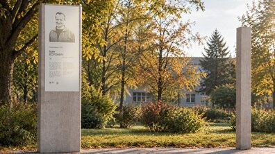 Візуалізація пам’ятної стели полковнику СБУ Івану Вороничу в парковому просторі. На вимощеній плиткою доріжці встановлено дві високі прямокутні кам’яні колони. На лівій колоні розміщена світла металева таблиця з портретом Івана Воронича, його ім’ям, званням і текстовою інформацією про нього. Друга колона не має видимих написів. Навколо ростуть дерева.