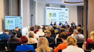 На фото відбувається резиденція. Учасники наповнили зал. На сцені сидять спікери, а позаду них – презентація