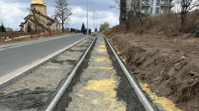 Ділянка дороги на вул. Віри, Надії, Любові. Уздовж проїжджої частини прокладено смугу майбутнього тротуару, обмежену свіжими бетонними бордюрами. Ліворуч проходить асфальтована дорога, якою їдуть автомобілі; обабіч встановлені ліхтарі.