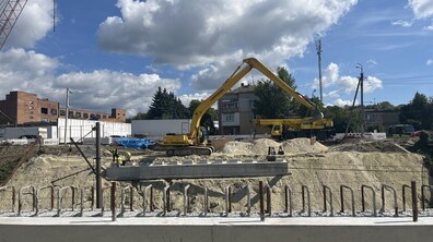 Майданчик будівництва шляхопроводу. На передньому плані видно масивну сіру бетонну балку з вертикальним рядом металевих арматурних стрижнів для подальшого бетонування. У центрі кадру на високому піщаному насипі працюють робітники у жилетах та стоїть великий жовтий гусеничний екскаватор із піднятою стрілою поруч із автокраном.