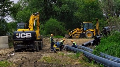 Працівники Львівводоканалу виконують заміну каналізаційного колектора. На розритій ділянці працює спецтехніка JCB, а монтажники готують до встановлення нові великі труби. Навколо — зелена лісиста місцевість.