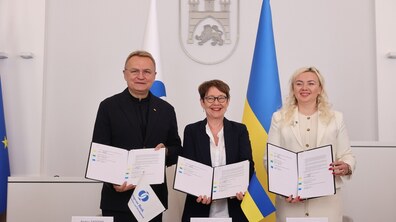 Мер Львова Андрій Садовий, Президентка Європейського банку реконструкції та розвитку Оділь Рено-Бассо та директорка ЛКП “Львівелектротранс” Ірина Іванишин тримають в руках підписаний меморандум про співпрацю та посміхаються у Великій сесійній залі Львівської міської ради. Позаду них видно прапори України та ЄБРР. По центру на стіні - Герб Львова