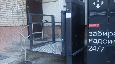 Вхід до під'їзду житлового будинку, обладнаний підйомником. Металева платформа підйомника з рифленою підлогою та сірими поручнями піднята на рівень вхідних дверей. Зліва від підйомника видно невеликі бетонні сходи з металевими перилами, що ведуть до темних вхідних дверей. Стіна будинку викладена зі світлої цегли.