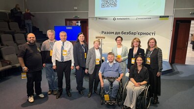Учасниками обговорення законопроєкту про формування безбар’єрного суспільства в Україні позують у залі на тлі екрана з написом «Разом до безбар'єрності». П'ятою зліва стоїть Ірина Маруняк у сірому піджаку та темних штанах. Усі учасники з бейджиками на шиї усміхаються та дивляться в камеру. На задньому плані також видно ряди сидінь амфітеатру та двох людей на сходах.