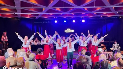 Сценічний виступ учасників, які відвідують заняття з танців у «Джерелі». Учасники, одягнені в українські народні костюми, стоять в один ряд із піднятими руками, завершуючи танець, а глядачі у залі аплодують їм. Сцену освітлюють яскраві синьо-фіолетові та помаранчеві прожектори.