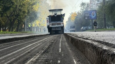 Демонтаж старого асфальтового покриття на вулиці Мазепи. У центрі кадру дорожня фреза знімає верхній шар асфальту. Обабіч дороги ростуть дерева, а вдалині видно працівників і транспорт.