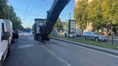У центрі стоїть велика фреза з піднятим конвеєром, яка знімає старе дорожнє покриття. Ліворуч видно білий фургон, попереду — тимчасові дорожні огорожі. На задньому плані є автобус, автомобілі, пішоходи, дерева та житлові будинки.