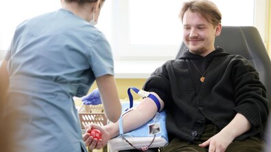 Усміхнений молодий чоловік здає кров, сидячи у медичному кріслі. Медсестра у блакитній формі готує пробірки для забору крові, поки донор тримає у руці червоний м'ячик-еспандер.