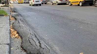 Вул. Зелена. Уздовж бордюру видно деформоване та потріскане асфальтне покриття з піднятими ділянками. На задньому плані рухаються автомобілі, а обабіч вулиці розташовані будівлі та припаркований транспорт
