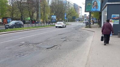 Проспект Чорновола. На передньому плані видно значні вибоїни та пошкодження асфальту в правій смузі руху. Дорогою їде білий автомобіль, праворуч тротуаром іде жінка. Уздовж вулиці ростуть дерева, розміщені рекламні щити та сучасні будівлі.