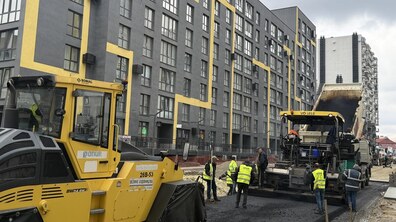 Процес укладання нового асфальту на вулиці Залізничній. На передньому плані працює велика дорожня техніка: жовтий каток і асфальтоукладальник, у який вантажівка висипає гарячий асфальт. Кілька робітників у світловідбивних жилетах розрівнюють покриття лопатами та контролюють процес. Ліворуч тягнеться багатоповерховий житловий будинок із сучасним фасадом у сірому кольорі з жовтими вертикальними акцентами та рядами вікон. Праворуч видно ще один житловий будинок і частину вулиці, де тривають роботи.