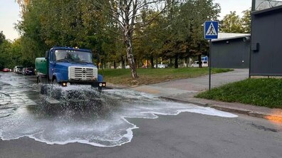 Поливальна машина їде міською вулицею. Попереду вантажівки видно широкий струмінь води, який розтікається по асфальту й утворює бризки. Обабіч дороги ростуть дерева, праворуч розташовані тротуар, пішохідний перехід і сучасна будівля.