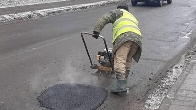 Ділянка міської дороги в холодну пору року — узбіччя та тротуар частково вкриті снігом. Посеред проїжджої частини видно темну круглу латану пляму — це свіже асфальтування. Поруч працівник комунальної служби. Він тримає перед собою ручну віброплиту — пристрій із металевою основою та ручкою, який ущільнює холодний асфальт і вирівнює поверхню дороги. На задньому плані видно автомобіль, що проїжджає повз місце ремонту.