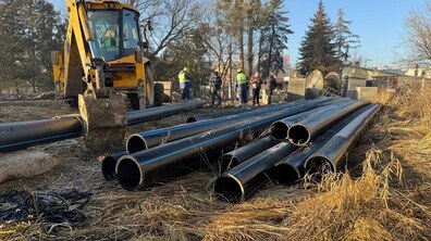 На передньому плані - довгі чорні пластикові труби великого діаметру, складені паралельно одна до одної. Ліворуч стоїть жовтий екскаватор-навантажувач. Його ківш опущений і торкається однієї з труб, ніби готується її пересунути або підняти. На задньому плані — кілька працівників Львівводоканалу у робочому одязі.