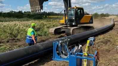 Роботи із заміни водогону в полі. На передньому плані лежить довга чорна поліетиленова труба великого діаметра, поруч встановлене обладнання для її зварювання. Біля труби стоїть працівник у сигнальному жилеті, а поряд працює гусеничний екскаватор із піднятим ковшем.