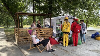 Локація «Літньої бібліотеки» у парку «Горіховий гай». Поряд із дерев'яним навісом, де дитина з дорослим граються конструктором, розташовані зона відпочинку з кріслами-мішками та намет з книжковими полицями. У центрі уваги — діти в яскравих жовтому та червоному костюмах з наплічниками, а на передньому плані — жінка відпочиває в шезлонгу за читанням книги.