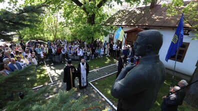 Подвір’я біля одноповерхового будинку музею. У центрі — вузька мощена доріжка, вздовж якої стоять двоє священників у чорному церковному вбранні. Навколо, півколом, зібралася велика кількість людей — дорослі й багато дітей, частина з них у вишиванках. Позаду видно будівлю музею з білими стінами і коричневим дахом. Поруч майорять два прапори: синьо-жовтий український і синій із жовтим тризубом. Фото архівне.