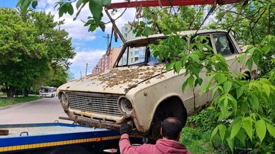 Евакуатор підіймає старий покинутий автомобіль світлого кольору, щоб завантажити його. Працівник скеровує процес евакуації авто з-під зелених дерев.