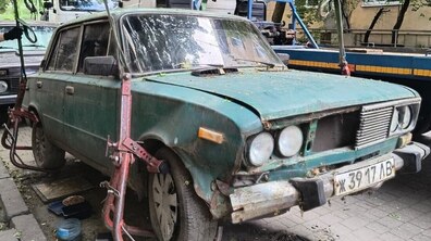 Припаркована на міській вулиці стара, іржава та брудна зелена автівка, вона має вм'ятини на капоті, розбиті фари та відсутні деталі бампера. Машину піднімають на евакуатор за допомогою спеціальних тросів, які зачеплені за колеса.