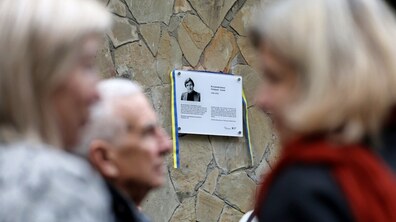 У центрі фото - розміщена на стіні будинку меморіальна інформаційна таблиця з чорно-білим портретом та біографічними відомостями про Володимира Івасюка. Табличку обрамляє стрічка у синьо-жовтих кольорах. Поруч стоять люди, які розмовляють між собою.