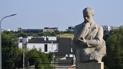 На передньому плані пам’ятник Тарасові Шевченку, який задумливо спирається рукою на підборіддя, а за ним простягається краєвид із житловими будинками, зеленими насадженнями та сучасною забудовою. На горизонті видно нові будівлі та відкриті зелені ділянки