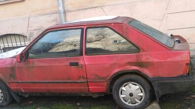 Старий занедбаний червоний легковий автомобіль стоїть узбіччі біля стіни будинку. По всьому його корпусу видно сліди іржі, бруду та зеленуватого нальоту.