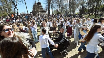 Великодні гаївки просто неба: люди в традиційному українському одязі танцюють і співають. В центрі — жінка і хлопчик танцюють разом із ветераном на кріслі колісному. На другому плані видно старовинну дерев’яну церкву та дерева.