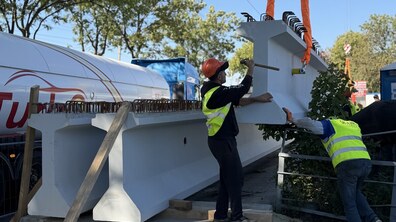 Процес розвантаження масивної сірої мостової балки за допомогою крана. Двоє робітників у яскраво-жовтих світловідбивальних жилетах і касках коригують її положення вручну, спрямовуючи до інших установлених балок.