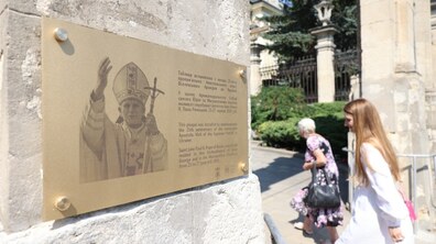 Меморіальна таблиця, прикріплена до кам'яної стіни. На ній вигравірувано портрет Папи Івана Павла ІІ та пам'ятний текст українською й англійською мовами. Поруч на розмитому тлі видно двох жінок і браму собору.