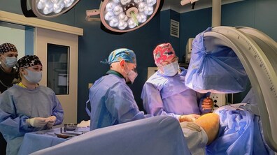В операційній команда лікарів проводить хірургічне втручання: медики у захисному одязі, масках і рукавичках зосереджено працюють біля пацієнта під яскравими операційними лампами. Поруч є сучасне операційне обладнання.