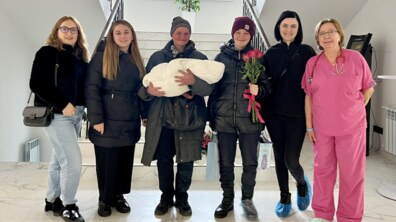 Анна з немовлям на руках, медичні працівниці та фахівці центру «Джерело» стоять у холі лікарні святої Анни: всі усміхнені та щасливі.