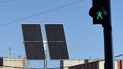 На тлі ясного блакитного неба поруч розташовані дві прямокутні сонячні панелі на опорі та пішохідний світлофор із увімкненим зеленим сигналом.