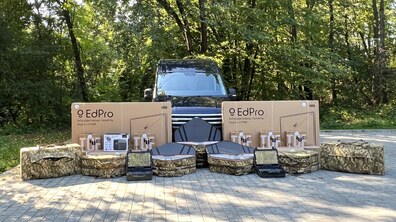 На відкритому брукованому майданчику стоїть темний автомобіль, а перед ними – багато коробок та кейсів з різним обладнанням, яке підготували для передачі військовим. Позаду видно зелені дерева.